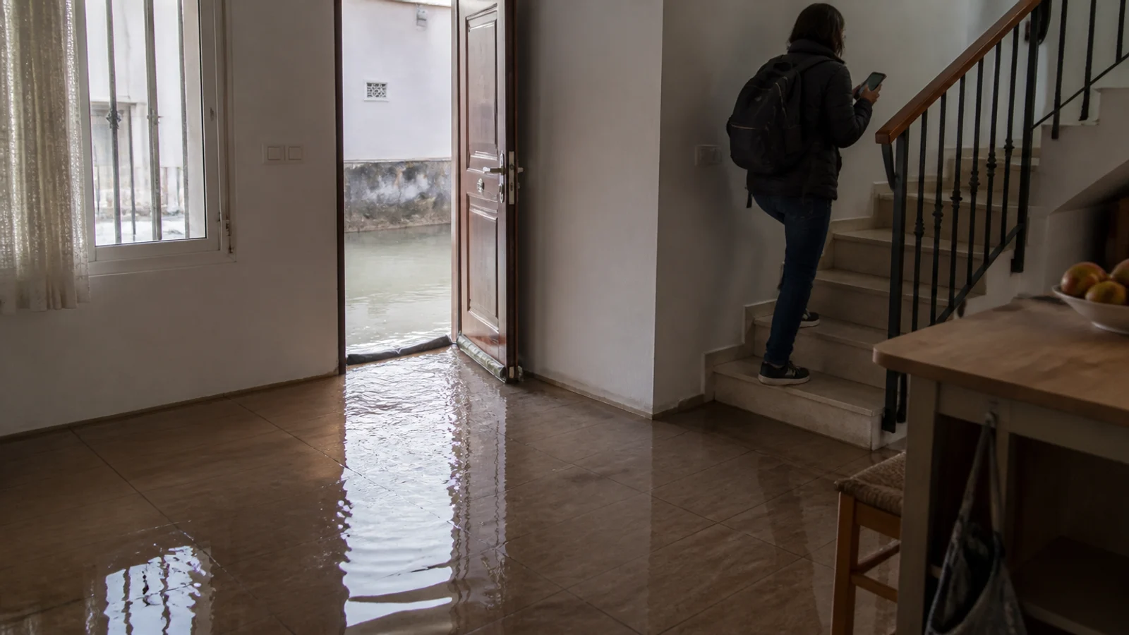 Agua turbia entrando bajo la puerta de una vivienda mientras una persona sube las escaleras hacia la planta alta
