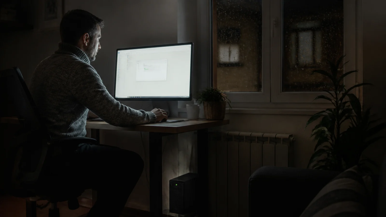 Persona guardando su trabajo en un ordenador iluminado a oscuras durante un apagón, con un SAI encendido bajo la mesa